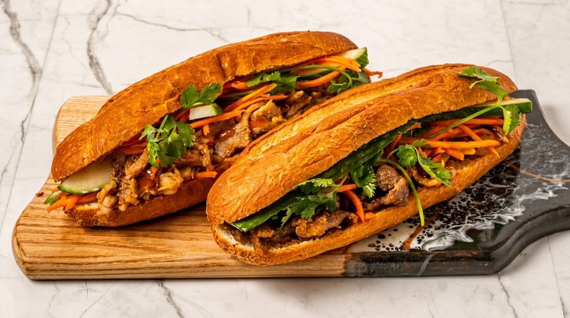 Two banh mi sandwiches with meat, pickled carrots, cucumber, and cilantro on a marble surface.