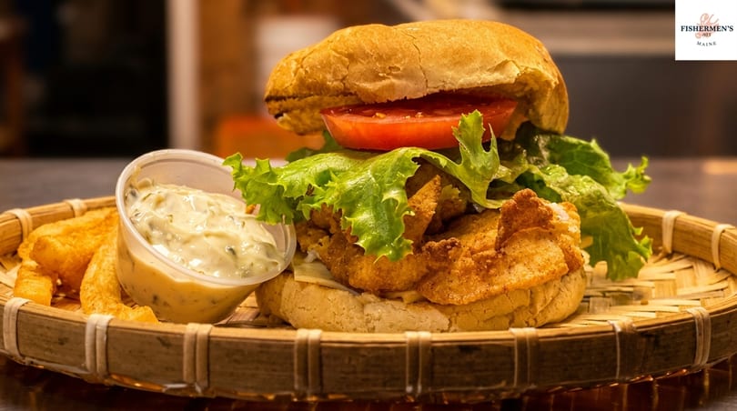 A fried fish sandwich with lettuce and tomato, served with fries and tartar sauce on a woven tray.
