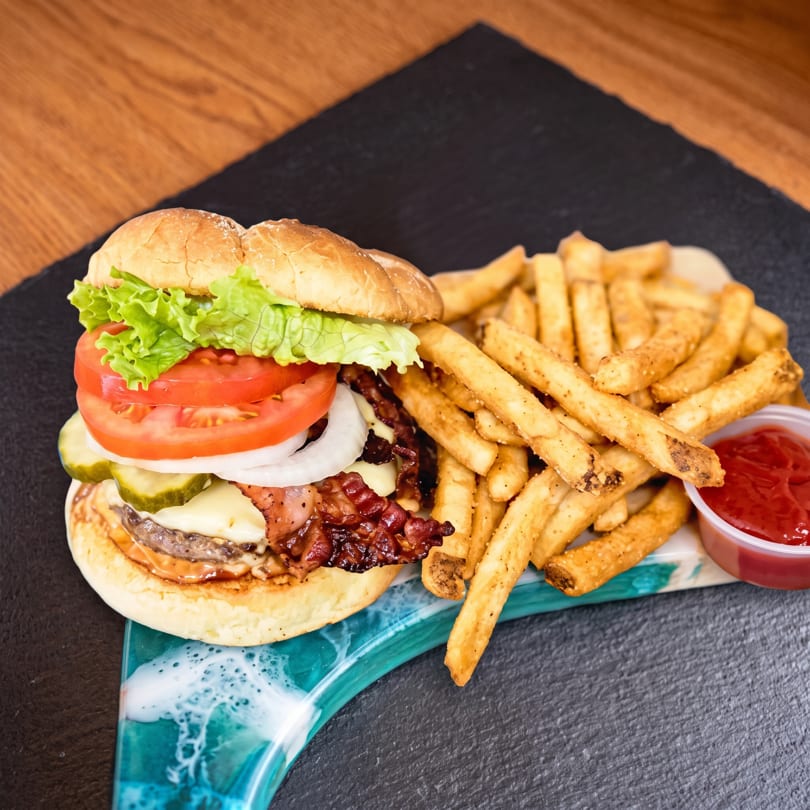 A loaded burger with bacon, cheese, lettuce, tomato, onion, and pickles, served with seasoned fries and ketchup.