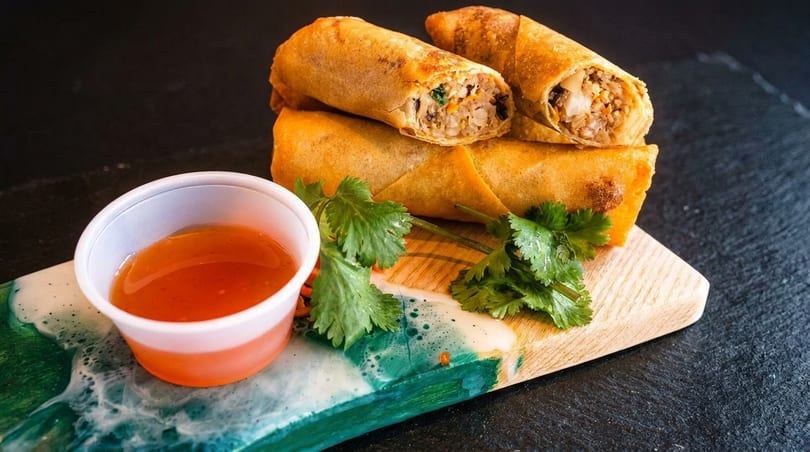 Crispy golden spring rolls, one cut open to show filling, with sweet chili sauce and cilantro.