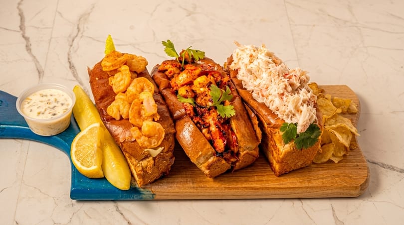 Three seafood rolls: shrimp, spicy shrimp, and crab, with chips, lemon, and sauce on a board.