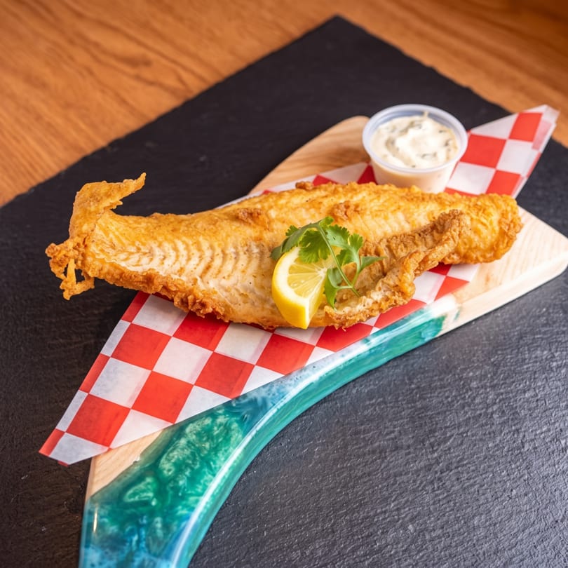Golden fried fish fillet with lemon, cilantro, and tartar sauce on a wooden board.